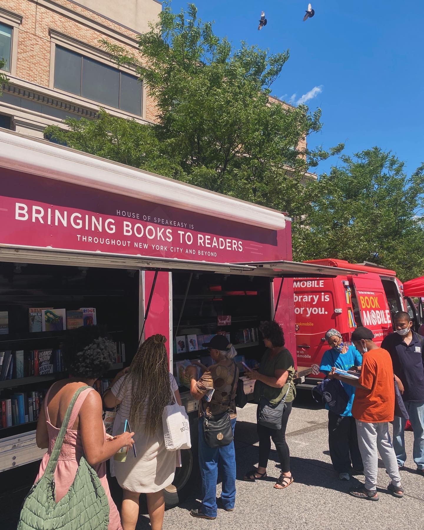 House of SpeakEasy's Bookmobile, parked on a sunny street behind a rack of bikes. A crowd of people is gathered by the Bookmobile's shelves, choosing their books.